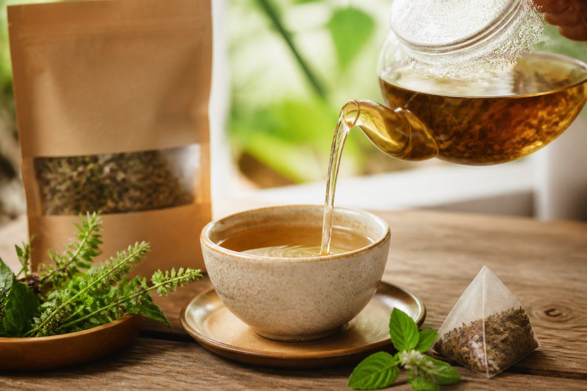 Ceramic cup of Ayurvedic herbal tisane poured beside a biodegradable pyramid tea bag and dried tulsi leaves