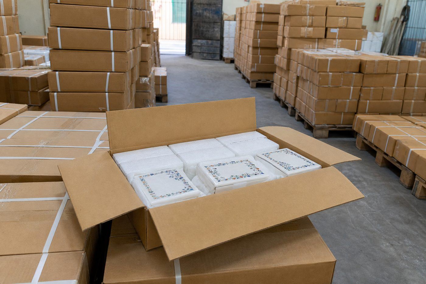 Palletized strapped cartons of marble inlay products in a dry Indian export warehouse with foam-protected jewelry boxes visible