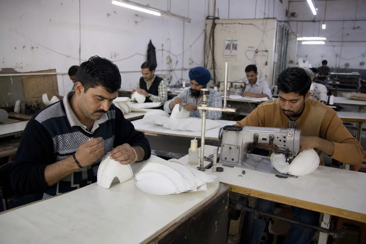 Workers hand-stitching and machine-stitching rugby ball panels on workbenches in a Jalandhar sports goods factory