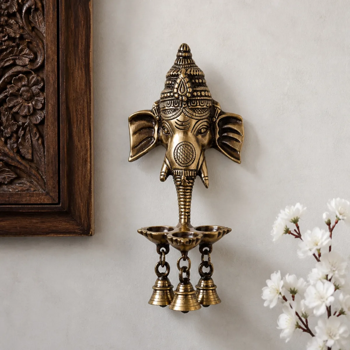 Handmade brass Ganesha deepak wall hanging with three bells on a grey plaster wall, antique gold from India
