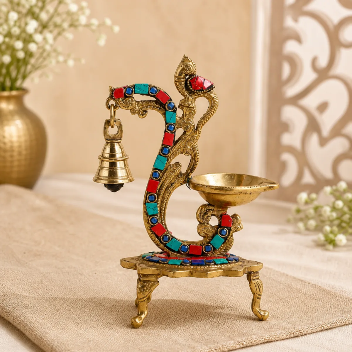 Handmade brass peacock diya with gemstone mosaic and hanging bell on linen, antique gold from India