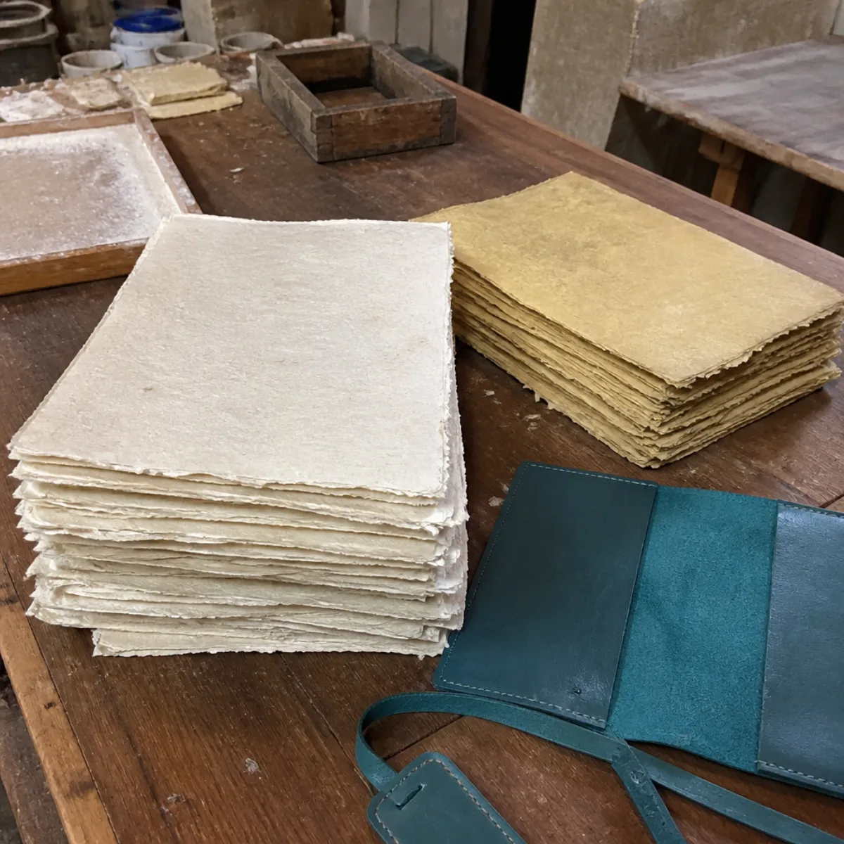 Handmade deckle-edge paper sheets and a teal leather cover on a workshop table