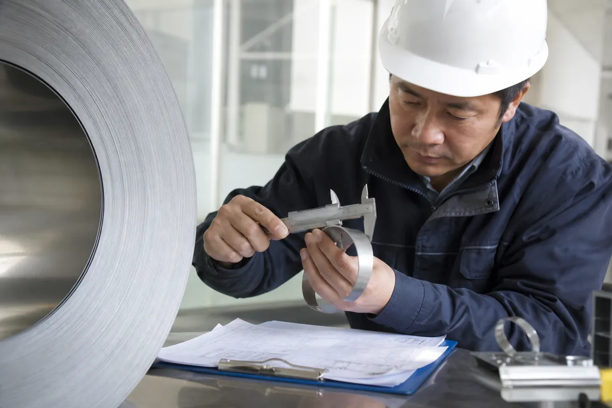Quality inspector reviewing an EN 10204 mill test certificate beside a stainless steel coil sample with calipers and hardness gauge before export release from India